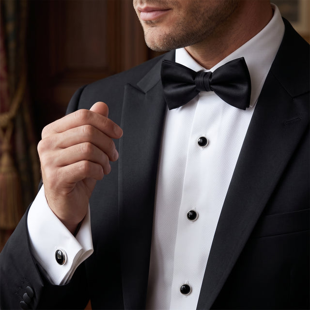 A man in a black tuxedo and bow tie, shown from mid-face to waist, adjusts his Tuxedo Shirt Studs and Cufflinks.