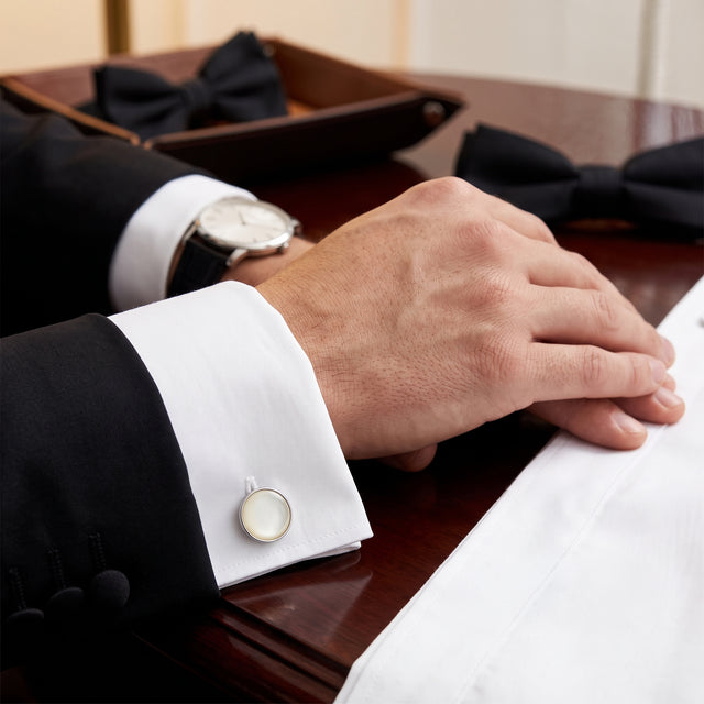 A man in a suit adjusts his Tuxedo Shirt Studs and Cufflinks on a crisp white shirt, with a watch and bow ties—classic accessories—neatly arranged on a wooden table.