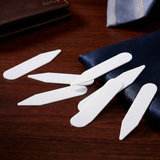 Six Collar Stays are arranged on a wooden surface beside a wallet and some navy blue fabric.