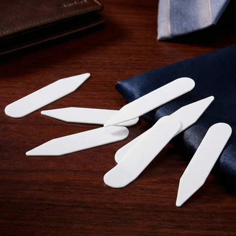 Six Collar Stays are arranged on a wooden surface beside a wallet and some navy blue fabric.