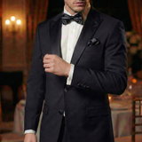 A man in a black tuxedo featuring Tuxedo Suit Buttons and a bow tie stands in an elegant, dimly lit room with a blurred background.