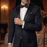 A man in a black tuxedo with Tuxedo Suit Buttons and a bow tie stands indoors at an elegant, dimly lit event.