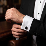 A man in a black tuxedo adjusts his white cuff, showcasing Tuxedo Shirt Studs and Cufflinks made of striking stainless steel at a bar.