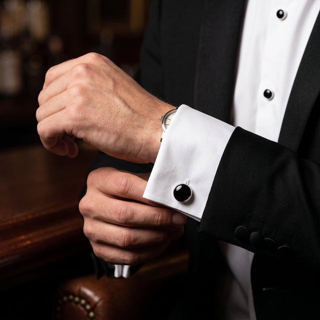A man in a black tuxedo adjusts his white cuff, showcasing Tuxedo Shirt Studs and Cufflinks made of striking stainless steel at a bar.