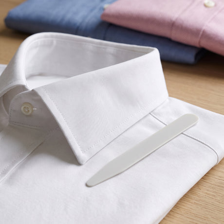 A neatly folded white dress shirt features a crisp look thanks to Collar Stays, with blue and pink shirts displayed in the background. Using Collar Stays helps the shirt maintain its sharp and polished appearance.